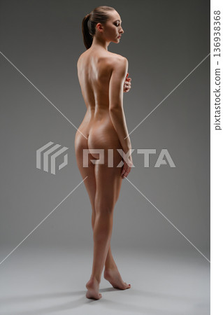 Elegant full-length nude woman posing from behind in studio. Athletic female body with graceful posture on a neutral gray background. Elegant full-length nude woman posing from behind in studio. Athletic female body with graceful posture on a neutral gray background. 136938368