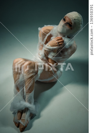 Conceptual portrait of a person wrapped in white gauze, sitting in a vulnerable pose. Conceptual portrait of a person wrapped in white gauze, sitting in a vulnerable pose. 136938391