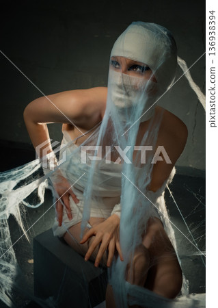 Spooky woman in mummy costume wrapped in white bandages and cobwebs against a dark background. 136938394