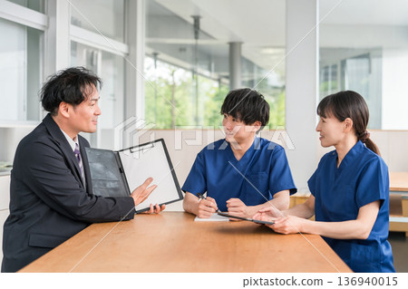 一位來自醫療設備製造商的男性商人和一位穿著手術服的男性和女性醫務人員正在進行銷售和商業談判 一位來自醫療設備製造商的男性商人和一位穿著手術服的男性和女性醫務人員正在進行銷售和商業談判 136940015