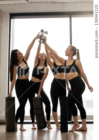 Four athletic women in matching black activewear raise water bottles in a celebratory toast after a fitness session, holding yoga mats Four athletic women in matching black activewear raise water bottles in a celebratory toast after a fitness session, holding yoga mats 136941220