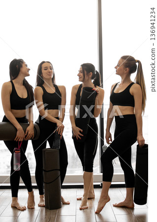 Four women in athletic wear stand together holding yoga mats and water bottles, ready for a workout session Four women in athletic wear stand together holding yoga mats and water bottles, ready for a workout session 136941234