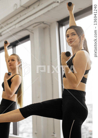 Two women in athletic wear are performing a workout routine using dumbbells in a bright studio setting 136941240