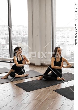 Two women in black activewear practice seated spinal twists on yoga mats in a bright studio with large windows overlooking a city Two women in black activewear practice seated spinal twists on yoga mats in a bright studio with large windows overlooking a city 136941294