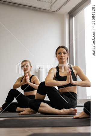 Two women in athletic wear are performing a seated spinal twist pose on yoga mats in a bright studio with large windows Two women in athletic wear are performing a seated spinal twist pose on yoga mats in a bright studio with large windows 136941295