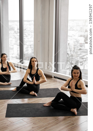 Three women in black activewear perform a seated spinal twist yoga pose on mats in a bright studio with large windows overlooking a city Three women in black activewear perform a seated spinal twist yoga pose on mats in a bright studio with large windows overlooking a city 136941297