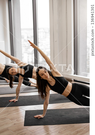 A group of women in black activewear perform side plank exercises on yoga mats in a bright studio with large windows 136941301
