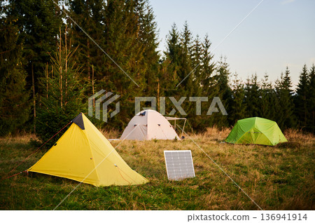 Solar panel in grassy field with tents in background highlights eco-friendly camping setup, surrounded by dense evergreen trees in evening. Integration of renewable energy in outdoor activities. Solar panel in grassy field with tents in background highlights eco-friendly camping setup, surrounded by dense evergreen trees in evening. Integration of renewable energy in outdoor activities. 136941914