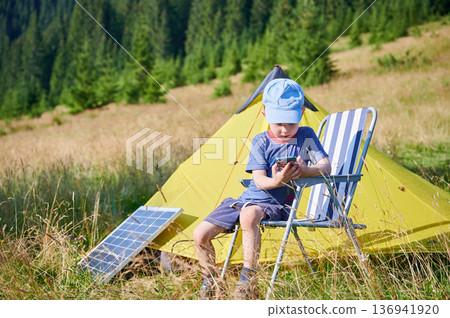Young child plays with smartphone while charging with photovoltaic solar panel near tourist tent in summer. Integration of renewable energy in outdoor camping activities. Young child plays with smartphone while charging with photovoltaic solar panel near tourist tent in summer. Integration of renewable energy in outdoor camping activities. 136941920