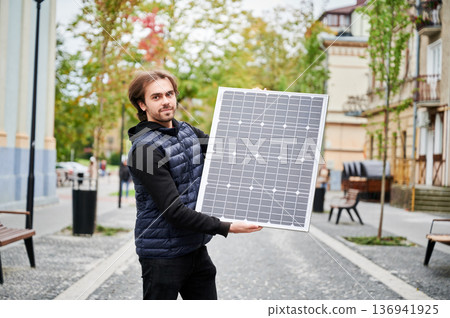 Portrait of man holding solar panel, stands in park. On background autumn trees and blurred city elements highlights integration of sustainable energy with everyday life. Portrait of man holding solar panel, stands in park. On background autumn trees and blurred city elements highlights integration of sustainable energy with everyday life. 136941925