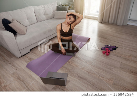 Woman practicing yoga at home on a purple mat in the living room while following an online video lesson during the afternoon Woman practicing yoga at home on a purple mat in the living room while following an online video lesson during the afternoon 136942584