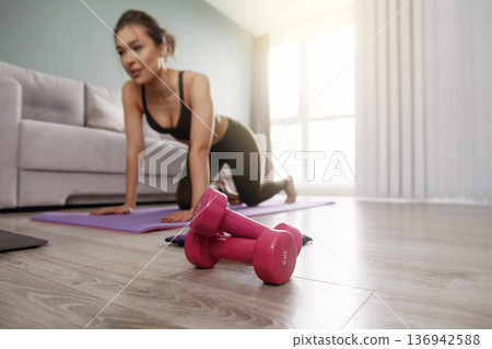 Woman exercises at home with dumbbells on a workout mat during the afternoon in a bright, airy living space Woman exercises at home with dumbbells on a workout mat during the afternoon in a bright, airy living space 136942588