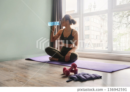 Woman sitting on yoga mat in workout attire drinking water and resting in a well-lit indoor space during her fitness routine Woman sitting on yoga mat in workout attire drinking water and resting in a well-lit indoor space during her fitness routine 136942591