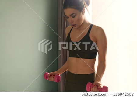 Woman exercises with pink dumbbells in a bright room during a fitness routine in the morning Woman exercises with pink dumbbells in a bright room during a fitness routine in the morning 136942598