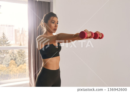 Woman performing strength training with pink dumbbells in a bright room while standing by large windows and enjoying the view Woman performing strength training with pink dumbbells in a bright room while standing by large windows and enjoying the view 136942600