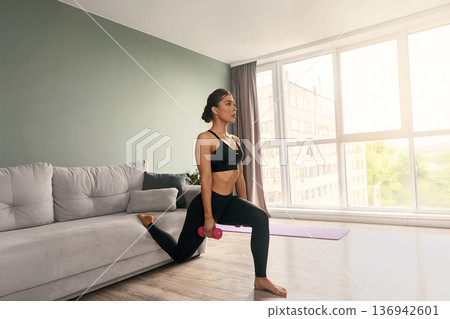 Fit woman doing lunges with dumbbell in modern living room during afternoon workout session 136942601