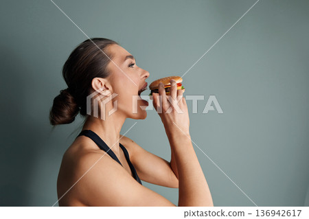 Woman enjoying a delicious burger against a simple green background while capturing a moment of tasty satisfaction 136942617