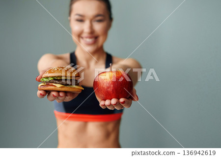 Woman holding an apple in one hand and a burger in the other while smiling against a simple background Woman holding an apple in one hand and a burger in the other while smiling against a simple background 136942619