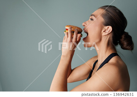 Woman enjoying a delicious burger against a simple green background while capturing a moment of tasty satisfaction 136942631