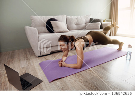 Woman doing a plank exercise at home while following an online workout on a laptop Woman doing a plank exercise at home while following an online workout on a laptop 136942636