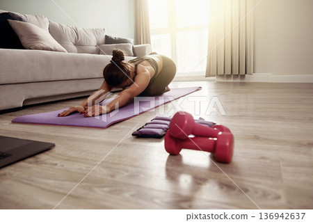 Woman stretch on yoga mat in living room with dumbbells and fitness equipment in morning light Woman stretch on yoga mat in living room with dumbbells and fitness equipment in morning light 136942637