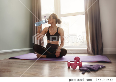 Woman sits on yoga mat in a bright room, drinking water after workout while weights are nearby Woman sits on yoga mat in a bright room, drinking water after workout while weights are nearby 136942638