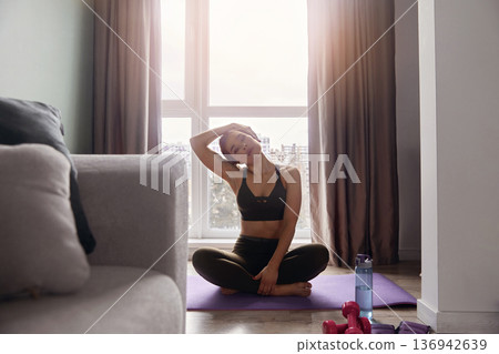 Woman stretches and practices yoga at home in the morning with weights and water bottle nearby Woman stretches and practices yoga at home in the morning with weights and water bottle nearby 136942639