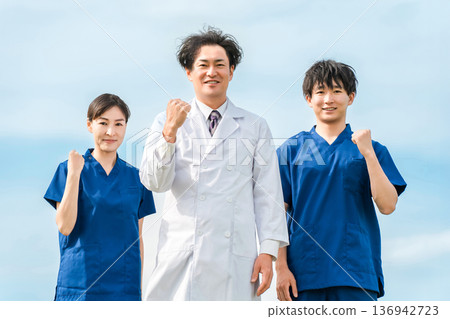 A male doctor in a white coat and male and female medical workers in scrubs under a blue sky A male doctor in a white coat and male and female medical workers in scrubs under a blue sky 136942723