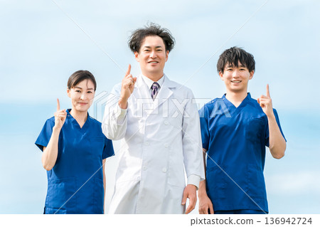 A male doctor in a white coat and male and female medical workers in scrubs under a blue sky 136942724