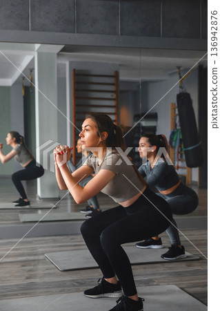 Women training in a gym while performing squats during a fitness session for a healthy lifestyle Women training in a gym while performing squats during a fitness session for a healthy lifestyle 136942876