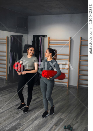 Two women carry yoga mats in a gym while talking after a fitness session Two women carry yoga mats in a gym while talking after a fitness session 136942888