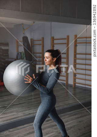 Woman trains with a fitness ball inside a gym during daylight hours Woman trains with a fitness ball inside a gym during daylight hours 136942892