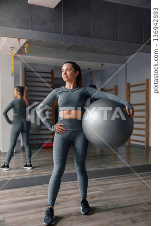 Woman stands with exercise ball in gym during training session for fitness and health Woman stands with exercise ball in gym during training session for fitness and health 136942893