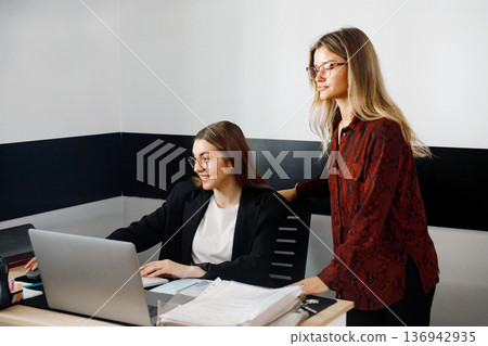 Women working together in an office during business hours at a desk with a laptop and paperwork 136942935