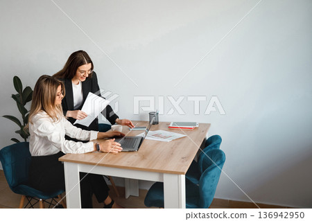Women discuss work at a table in an office during the day while using a laptop and reviewing papers Women discuss work at a table in an office during the day while using a laptop and reviewing papers 136942950