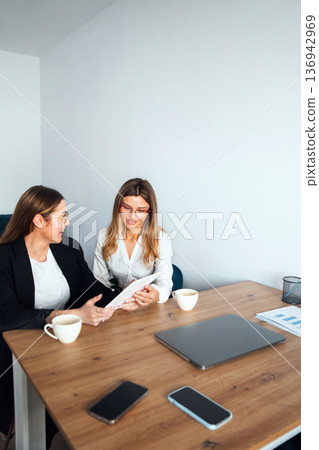 Women working together in an office discussing ideas during a meeting at a wooden table with laptops and coffee cups Women working together in an office discussing ideas during a meeting at a wooden table with laptops and coffee cups 136942969