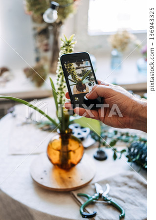 Hand holds smartphone to photograph floral arrangement in glass vase on table. influencer lifestyle, floral blogging, behind the scenes, mobile photography, sharing hobbies online 136943325