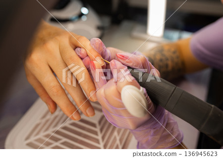 Close up of professional manicure procedure using electric nail drill. Nail technician wearing gloves treating female nails in beauty salon. Nail care, hygiene, cosmetic treatment concept. Close up of professional manicure procedure using electric nail drill. Nail technician wearing gloves treating female nails in beauty salon. Nail care, hygiene, cosmetic treatment concept. 136945623