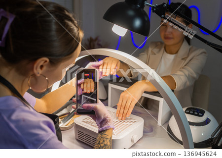Nail technician photographing finished manicure on female hands using smartphone under ring lamp in beauty salon. Professional nail care, content creation, social media and manicure service concept. 136945624