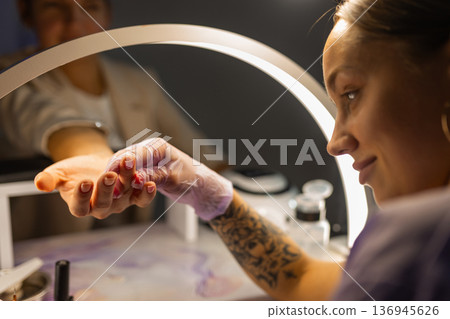 Professional nail technician performing manicure for female client under lamp in beauty salon. Nail care service, cosmetic treatment, hygiene, modern manicure workspace concept. 136945626