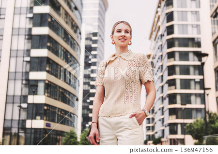Beautiful confident woman standing in city street with modern skyscrapers building in background. Stylish female model in urban environment and contemporary architecture outdoors Beautiful confident woman standing in city street with modern skyscrapers building in background. Stylish female model in urban environment and contemporary architecture outdoors 136947156