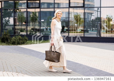 Portrait stylish young woman walking outdoors in city near modern building. Female wearing elegant beige summer outfit Sleeveless maxi dress, headscarf, flat shoes and large handbag, urban lifestyle Portrait stylish young woman walking outdoors in city near modern building. Female wearing elegant beige summer outfit Sleeveless maxi dress, headscarf, flat shoes and large handbag, urban lifestyle 136947161