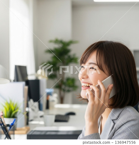 一位年輕的女員工在辦公室裡面帶著微笑地用智慧型手機通話。 136947556