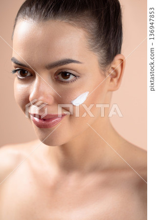 Beautiful woman applying white cream with fingers on cheek on the beige background. Portrait of beauty model with natural nude make up. Spa, skincare, wellness. Close up 136947853