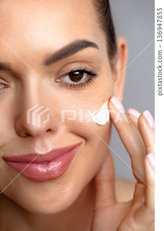 Beauty Woman face skin care. Beautiful close up portrait of a young woman with a glowing skin is applying a skincare product. Cream smear 136947855