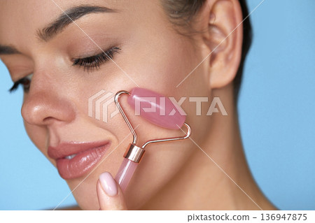 Woman using jade facial roller for face massage. Girl using natural massager tool for skin care and spa beauty treatment closeup portrait. 136947875