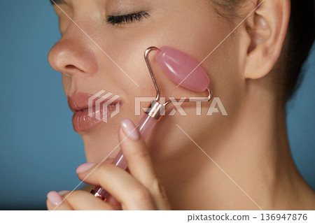Woman using natural pink quartz face roller on blue background, closeup. Face massage.. Woman using natural pink quartz face roller on blue background, closeup. Face massage.. 136947876