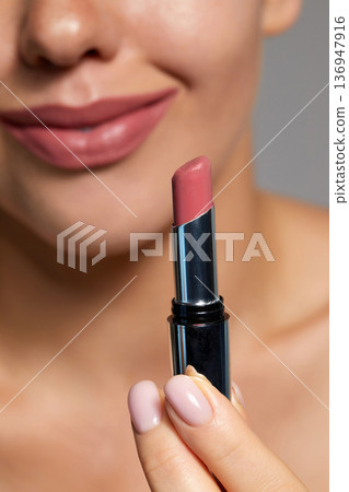 Woman applies nude lipstick while smiling in a skincare routine at a cosmetic studio with bright lighting 136947916