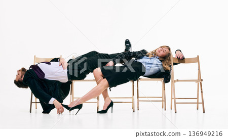 Two business professionals dramatically lying across wooden chairs in white studio. 136949216