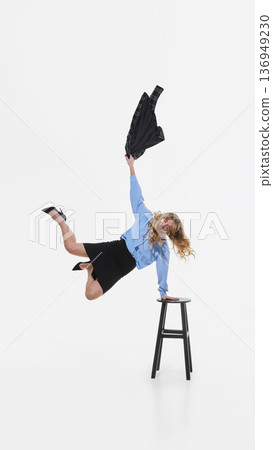 Young business woman balancing on stool while holding jacket mid air on white background. 136949230